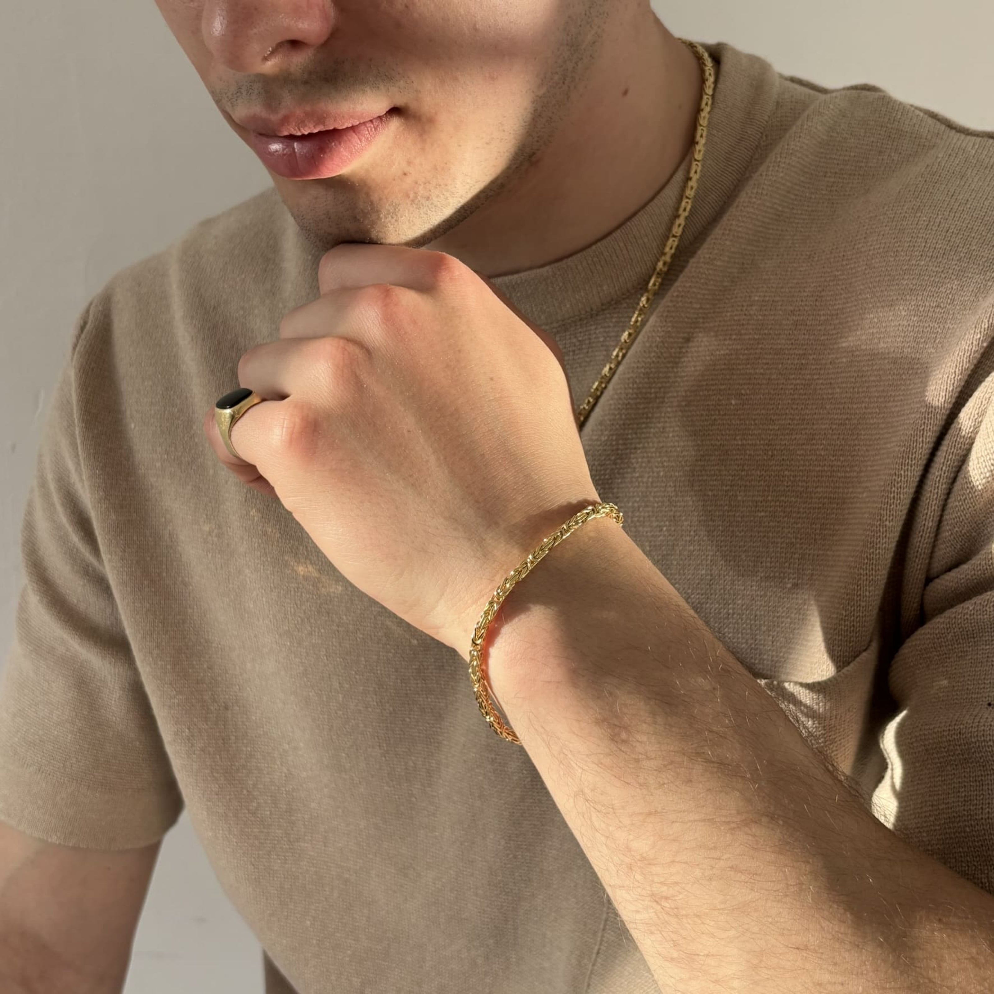 Mann mit beigefarbenem Shirt zeigt ein vergoldetes Königsarmband aus 925 Sterling Silber am Handgelenk – diamantiert veredelt und kombiniert mit einem Siegelring und einer goldfarbenen Halskette, präsentiert von Gold Dittrich.
