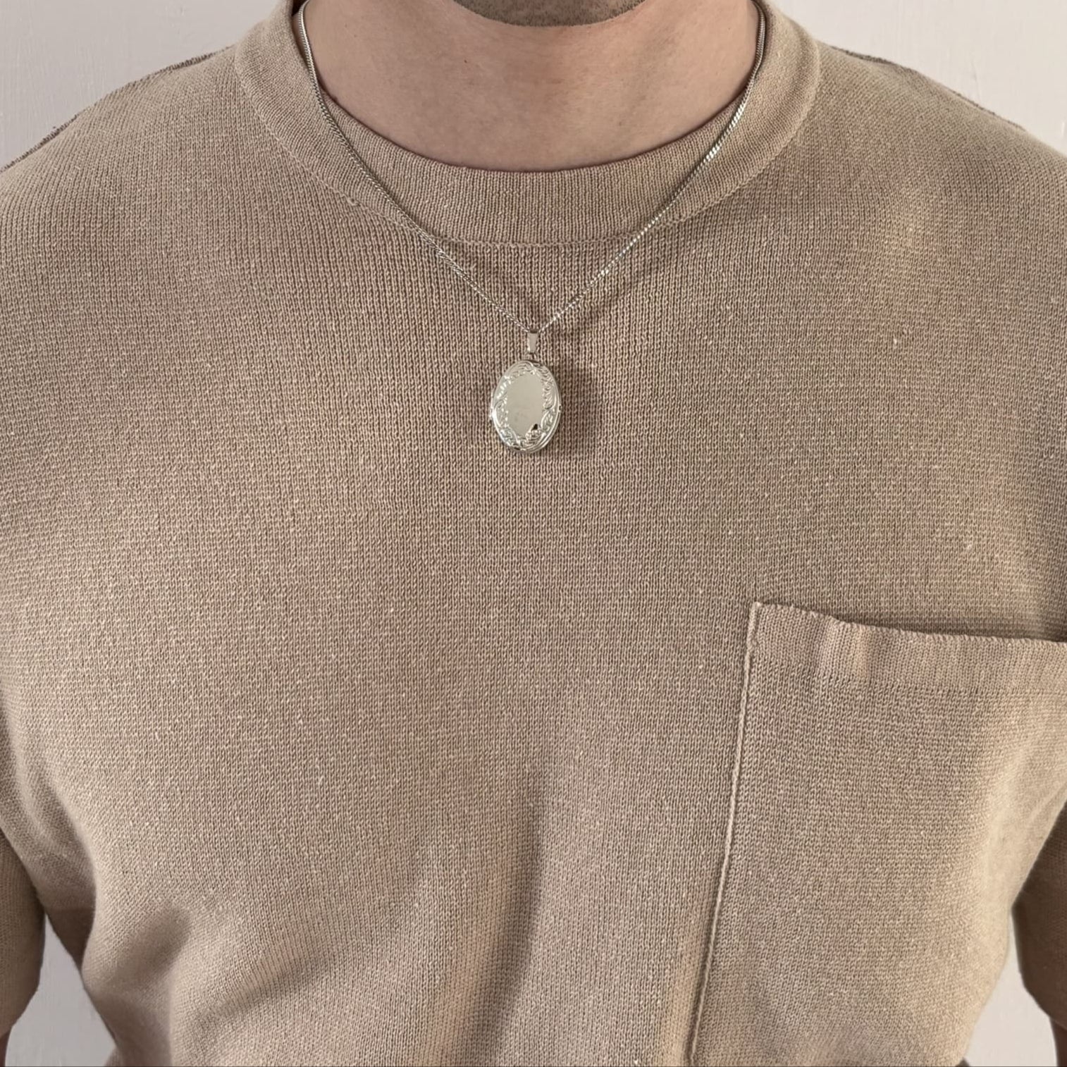 Person mit beigefarbenem Shirt trägt eine silberne Medaillon-Kette aus rhodiniertem 925 Sterlingsilber – ovaler Anhänger mit feinen Gravurdetails und Panzerkette, ideal zur Aufbewahrung von bis zu vier Fotos.